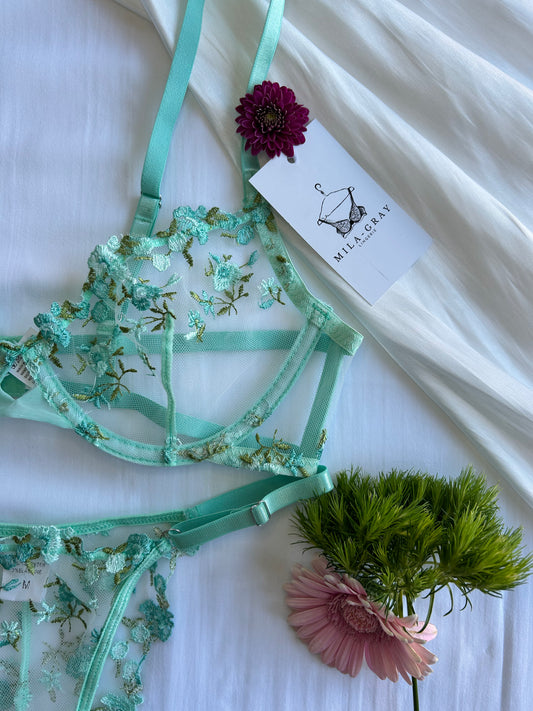 A set of lingerie featuring a underwire bra and matching garter belt with floral lace detailing, displayed on a white background with a 'Wild Poppy' brand tag and some floral decor.