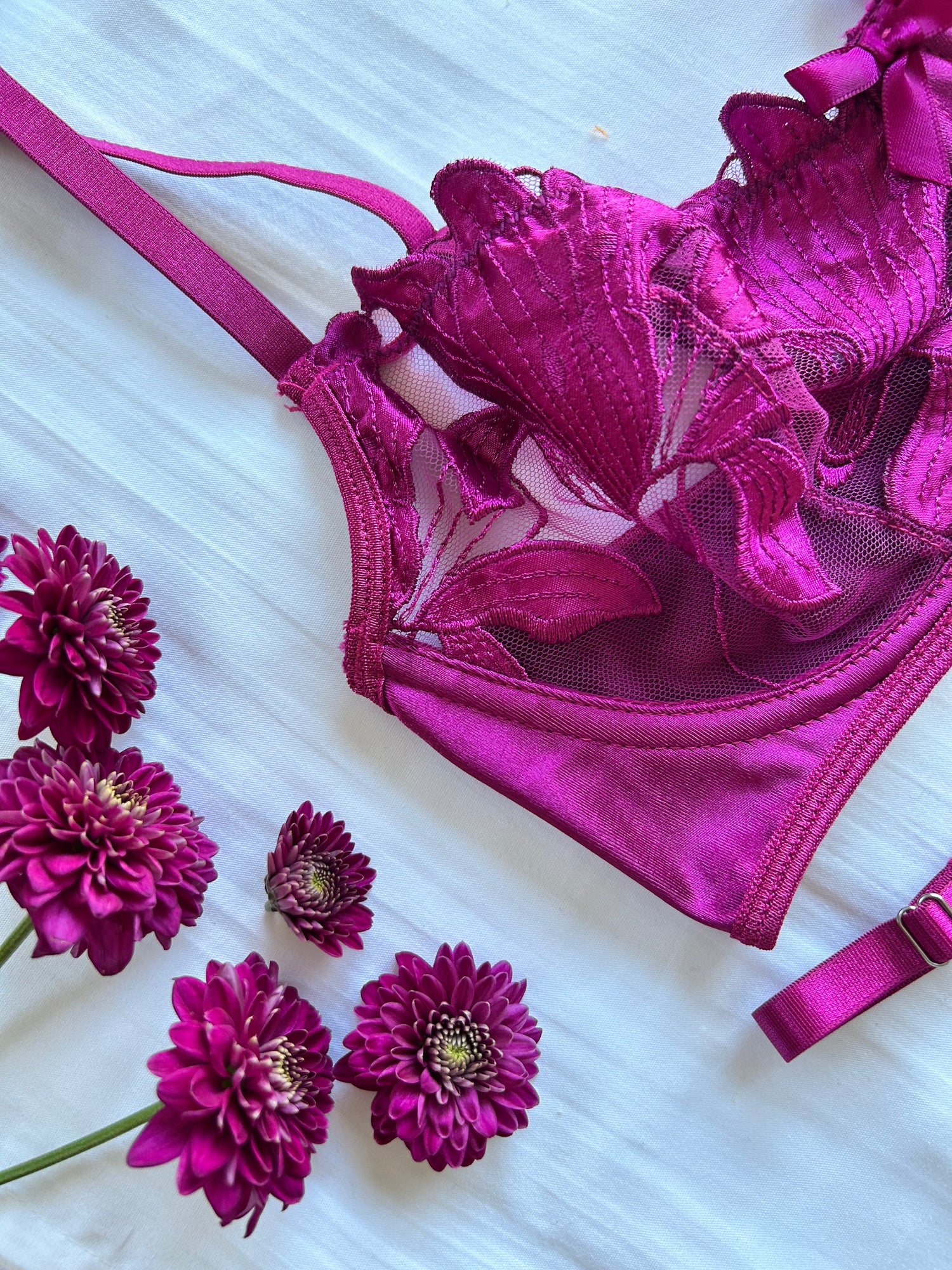 A wine red/purple lingerie set with adjustable straps, underwire support, and a ribbon bow accent, displayed on a white background surrounded by matching purple flowers.