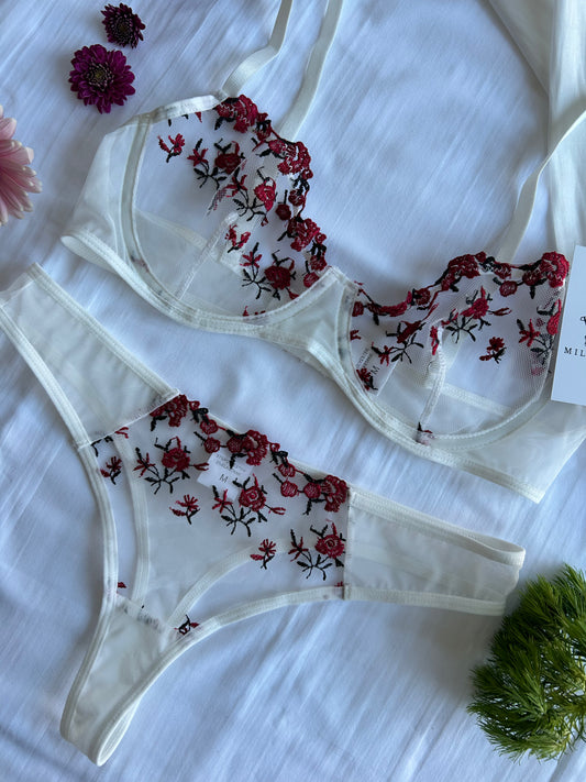 A white lingerie set with red floral embroidery lace, consisting of a bra and matching panties, displayed on a light background with floral accents.