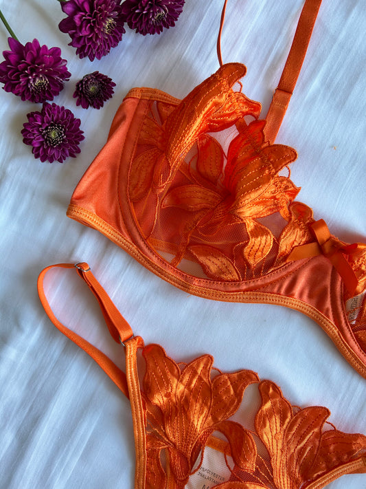 An orange lingerie set with a floral pattern, consisting of a underwire bra and a thong, displayed on a white surface with a bow detail. Some purple flowers are also visible in the corner.
